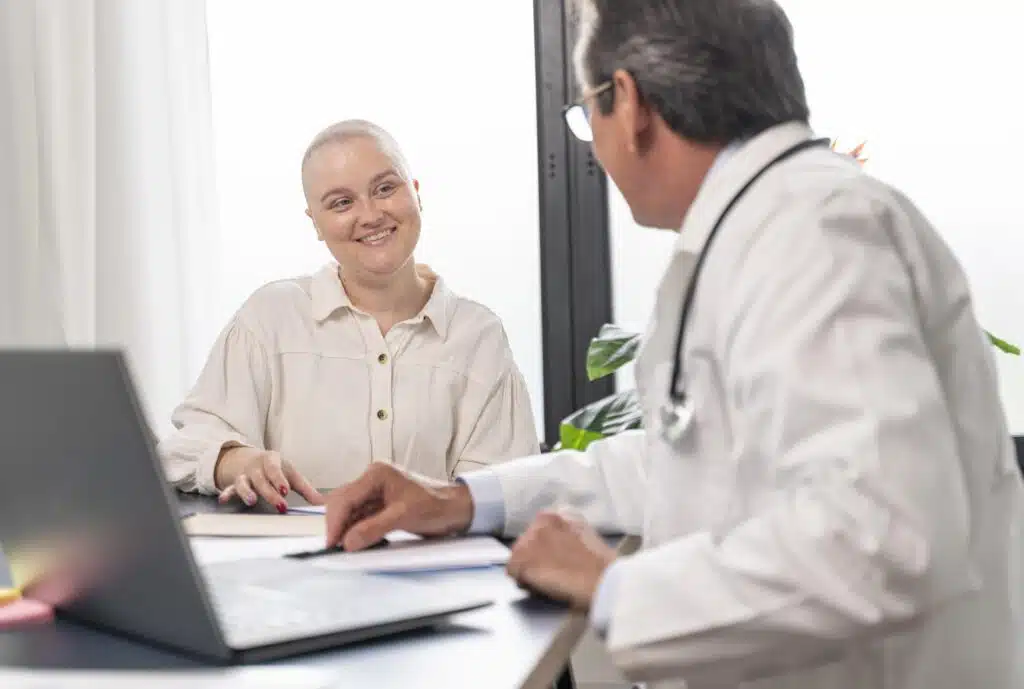 A woman with a short, shaved hairstyle, possibly indicative of undergoing chemotherapy, is smiling and conversing with her male doctor, who is dressed in a white coat with a stethoscope around his neck. They are both seated at a desk with a laptop, papers, and what appears to be a medical chart, suggesting a friendly consultation or discussion of the woman's treatment plan in a clinical setting.
